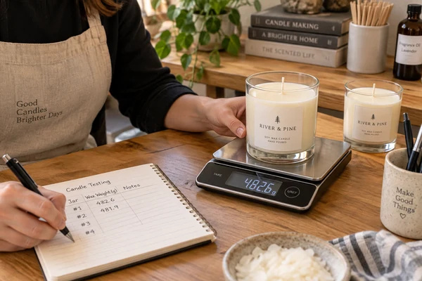 A person using a digital scale to weigh a candle jar filled with wax, with a notebook nearby for recording data