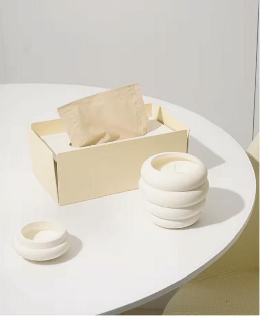 Ivory ceramic candle jar beside its open presentation box Pale presentation box beside a small ribbed vessel and round lid.
