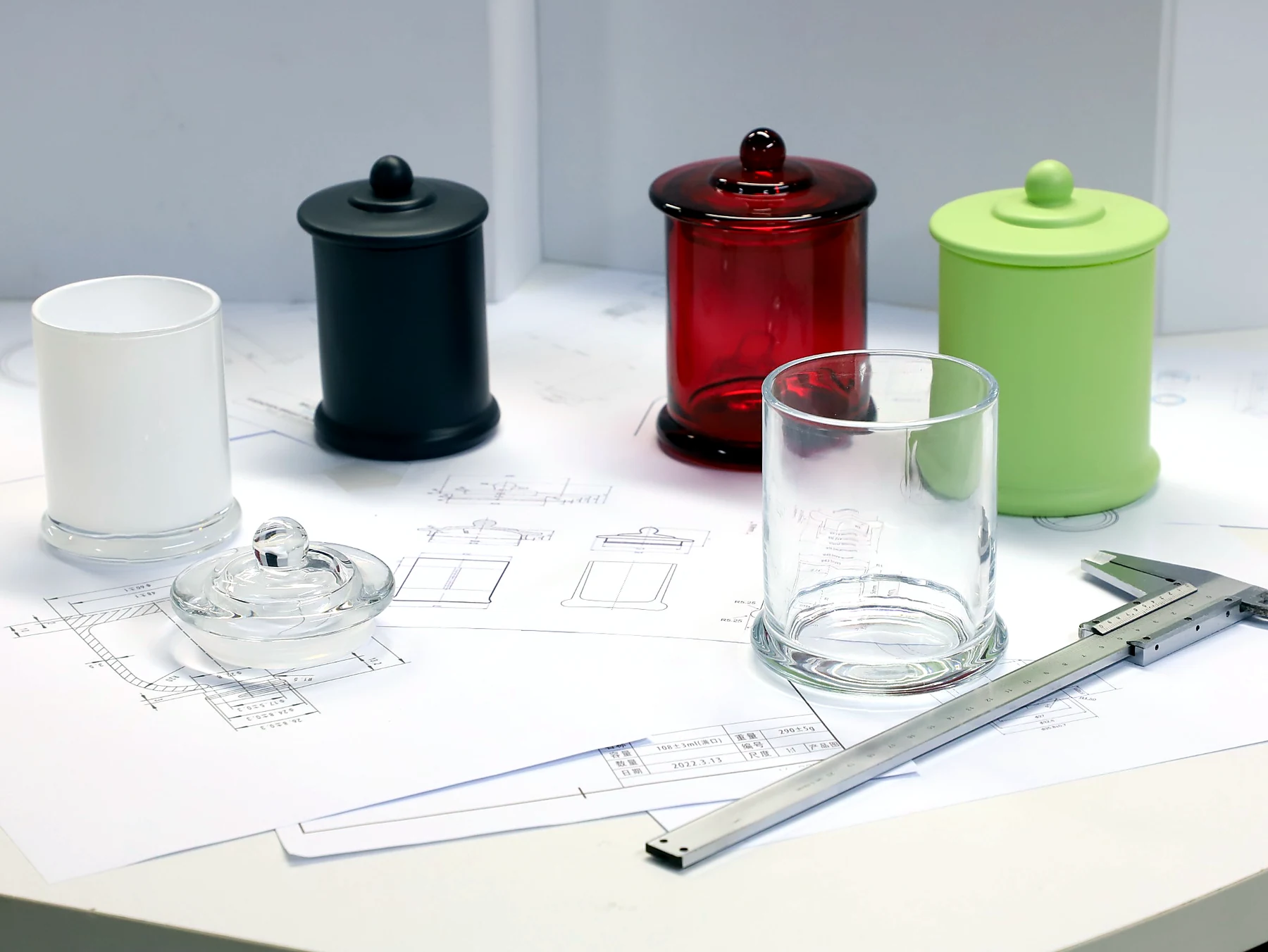 Finished glass candle jars in clear, white, black, red and green beside technical drawings and a caliper for physical finish approval